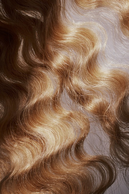 Close-up of wavy blonde human hair. The hair has a golden, dimensional color and a soft wave pattern, filling the frame.