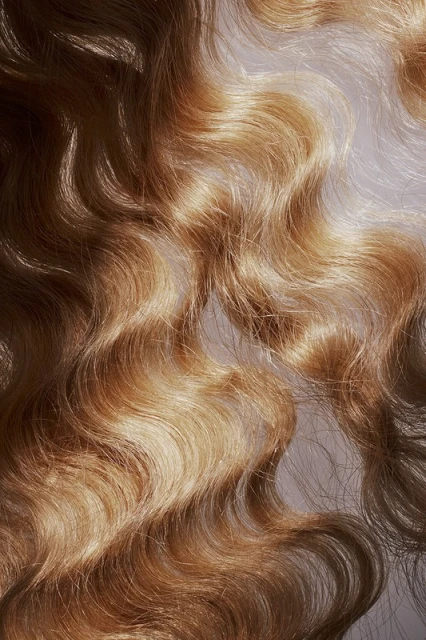 Close-up of wavy blonde human hair. The hair has a golden, dimensional color and a soft wave pattern, filling the frame.