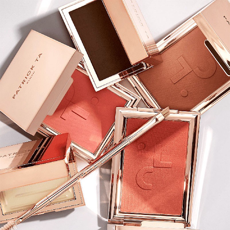 Patrick Ta blush compacts and brush displayed in a beauty still life. The composition highlights warm tones and cosmetic textures.
