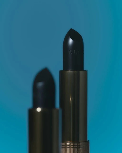 Close-up studio shot of two tubes of dark lipstick, one in focus and one blurred, against a gradient light blue background.