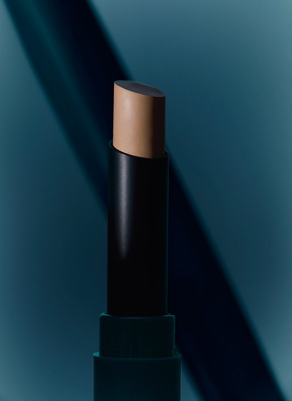 Close-up shot of a makeup concealer stick with a matte black tube and tan color makeup, against a blue background with dramatic lighting.