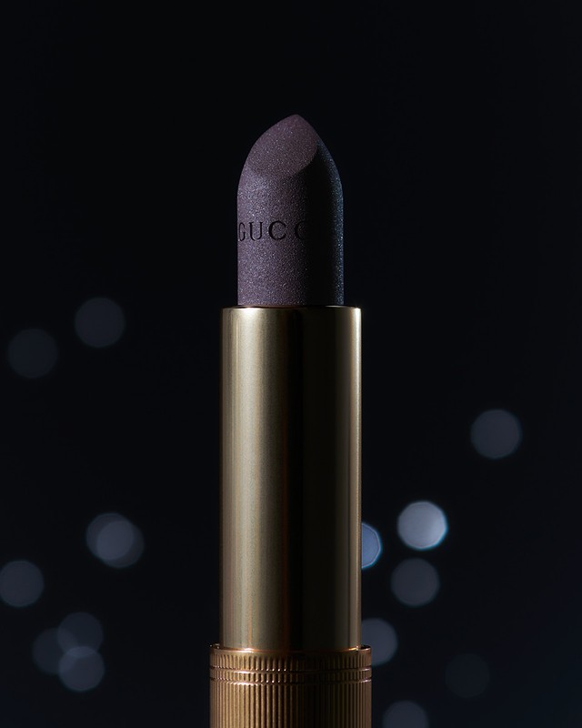 Close-up studio shot of Gucci lipstick. The product has a golden tube and a purple bullet with the brand name visible on the bullet.