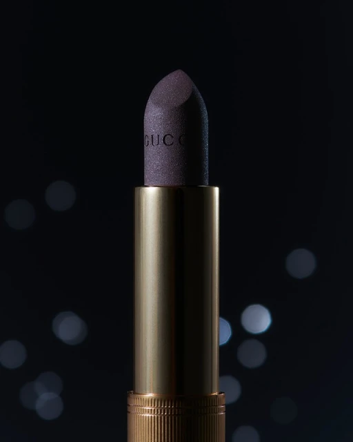 Close-up studio shot of Gucci lipstick. The product has a golden tube and a purple bullet with the brand name visible on the bullet.