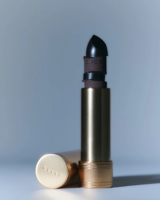 Still life shot of opened black lipstick in a gold tube, next to the lipstick cover with Gucci logo, on a pale blue surface.