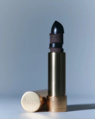 Still life shot of opened black lipstick in a gold tube, next to the lipstick cover with Gucci logo, on a pale blue surface.