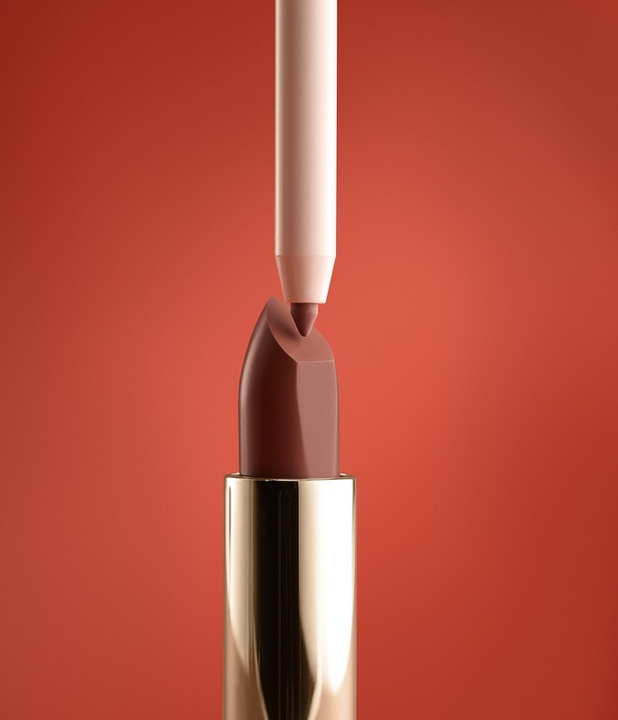 Close-up shot of brown lipstick with gold tube and nude-colored lip liner pointed at the tip, set against a reddish-brown background.