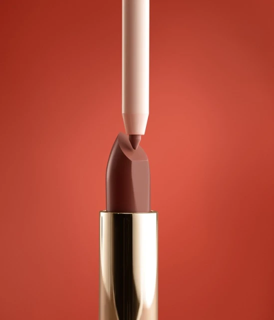 Close-up shot of brown lipstick with gold tube and nude-colored lip liner pointed at the tip, set against a reddish-brown background.