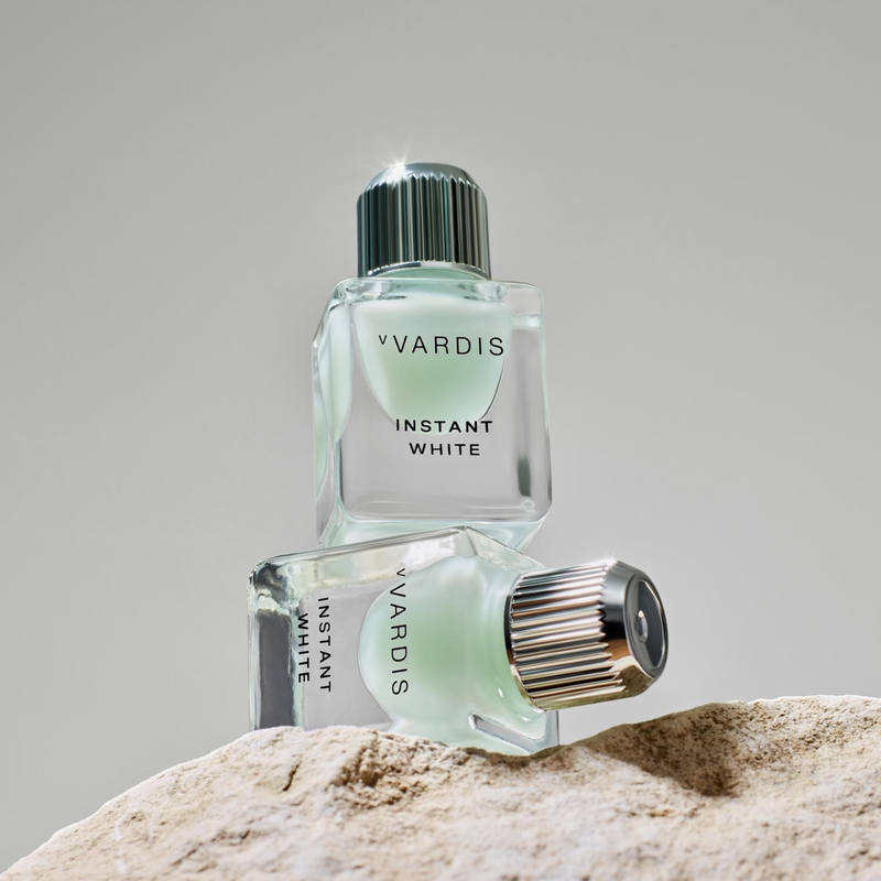 Two clear glass bottles of Vvardis Instant White sit stacked on a textured stone surface against a soft grey background, studio shot.