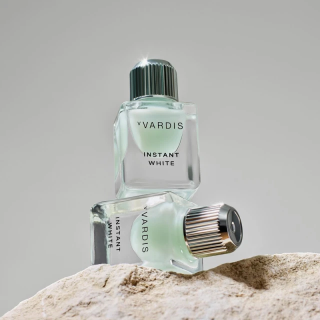 Two clear glass bottles of Vvardis Instant White sit stacked on a textured stone surface against a soft grey background, studio shot.
