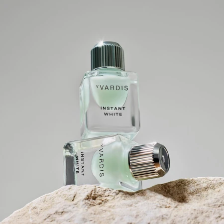 Two clear glass bottles of Vvardis Instant White sit stacked on a textured stone surface against a soft grey background, studio shot.