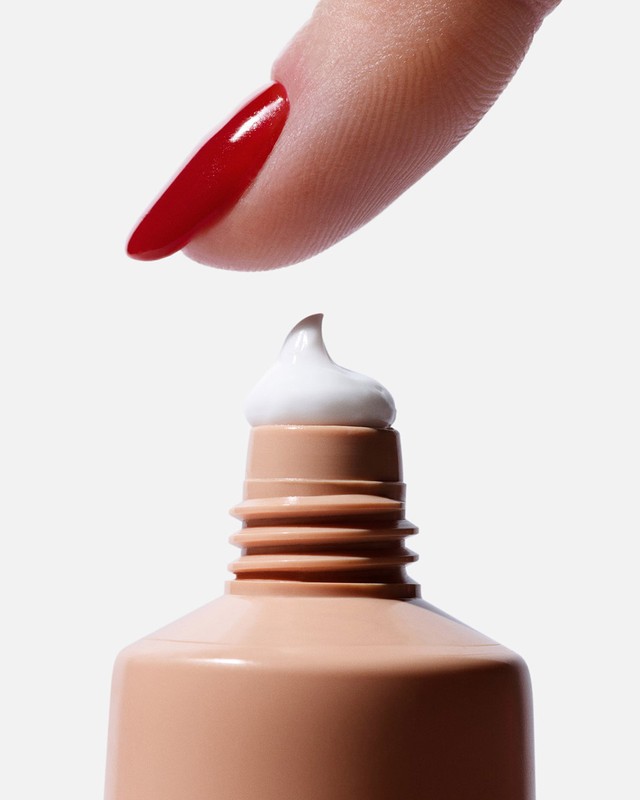 Close-up shot featuring a manicured finger touching a dab of white cream on a tan tube against a clean white background.