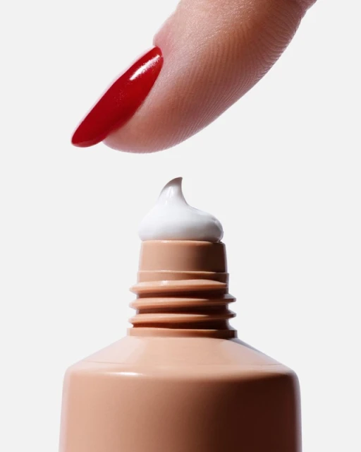 Close-up shot featuring a manicured finger touching a dab of white cream on a tan tube against a clean white background.