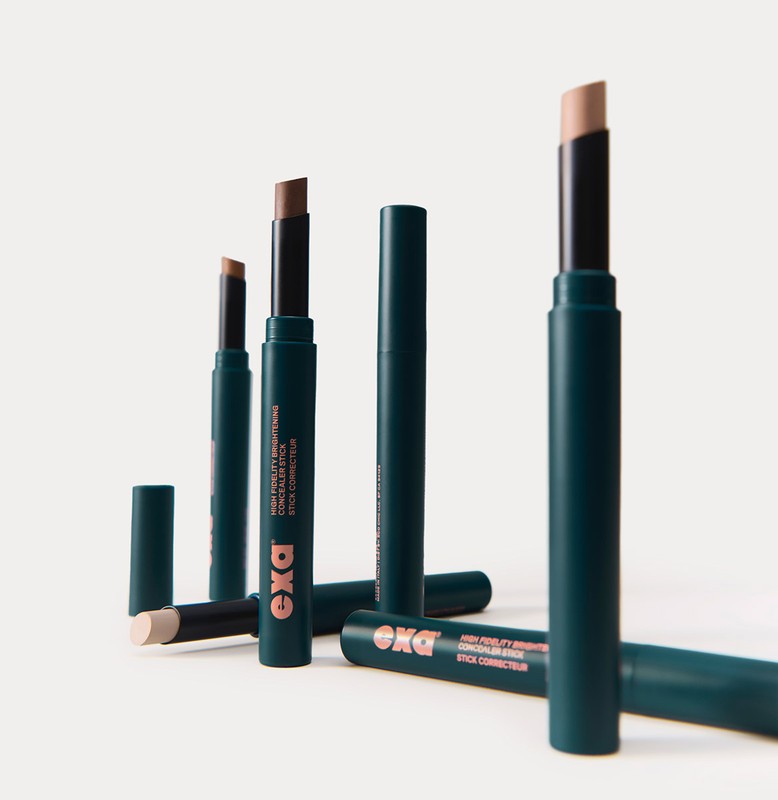 Several teal concealer sticks by EXA Cosmetics stand against a plain background, some with caps off, showing tan and brown colors.