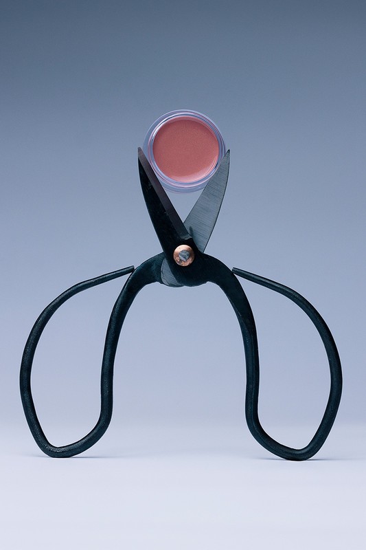 Beauty product in clear container held by black vintage scissors against a light gray backdrop, emphasizing beauty and vintage style.