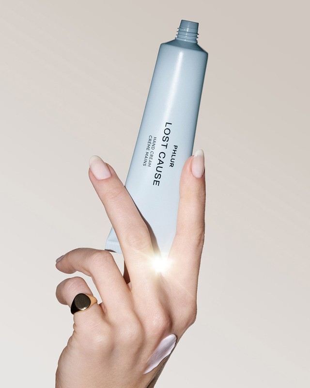 A hand model applies PHLUR Lost Cause hand cream from a light blue tube, adorned with a gold ring, against a beige backdrop.