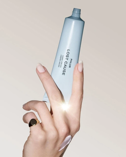 A hand model applies PHLUR Lost Cause hand cream from a light blue tube, adorned with a gold ring, against a beige backdrop.