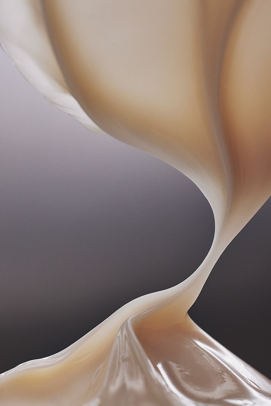 Abstract close-up of viscous, beige fluid with a glossy sheen. Gradated gray background creates contrast with the swirling form.