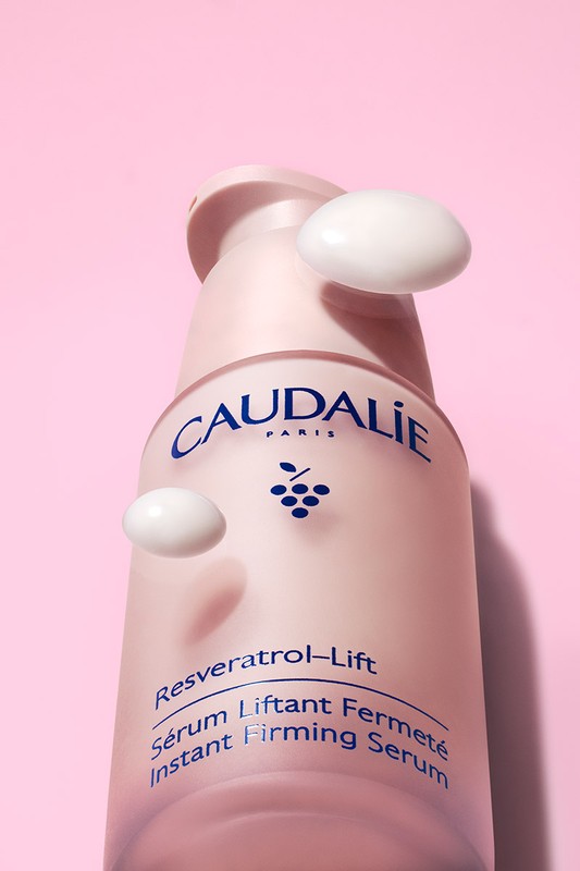 Close-up of Caudalie Resveratrol Lift instant firming serum against a light pink background, with visible drops of white serum.