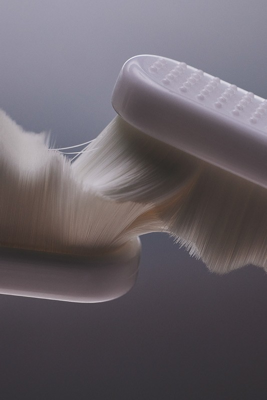Close-up studio shot of a white, soft-bristled toothbrush head against a grey gradient background, highlighting the fine bristles and texture.
