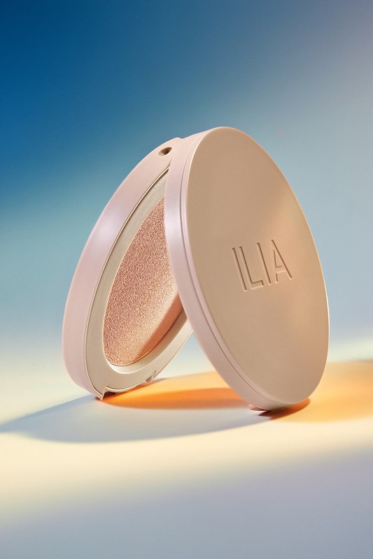 Close-up of a makeup compact featuring a powder product with the brand name 'ILIA' visible on the case, set against a blue background.
