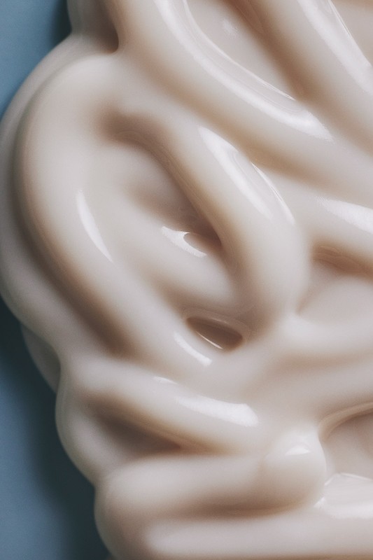 Close-up of thick, swirled, glossy beige cream against a light blue backdrop, showcasing smooth texture and reflective surface.