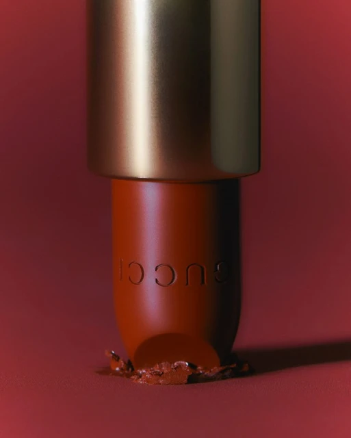 Close-up shot of a broken orange-red Gucci lipstick tube against a matching red background. Luxurious makeup with a gold lid.