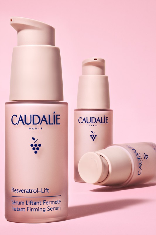 Three Caudalie Resveratrol-Lift serum bottles in varying sizes and orientations against a pink backdrop in a studio setting.
