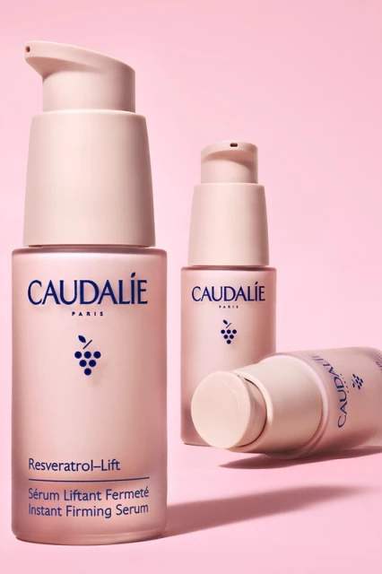 Three Caudalie Resveratrol-Lift serum bottles in varying sizes and orientations against a pink backdrop in a studio setting.