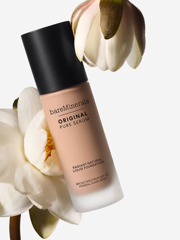BareMinerals Original Pure Serum foundation bottle close-up with a black cap, surrounded by white water lily flowers against a white backdrop.