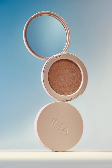 Elevated view of tan Ilia makeup compacts on a blue gradient backdrop with soft light, highlighting beauty product details.