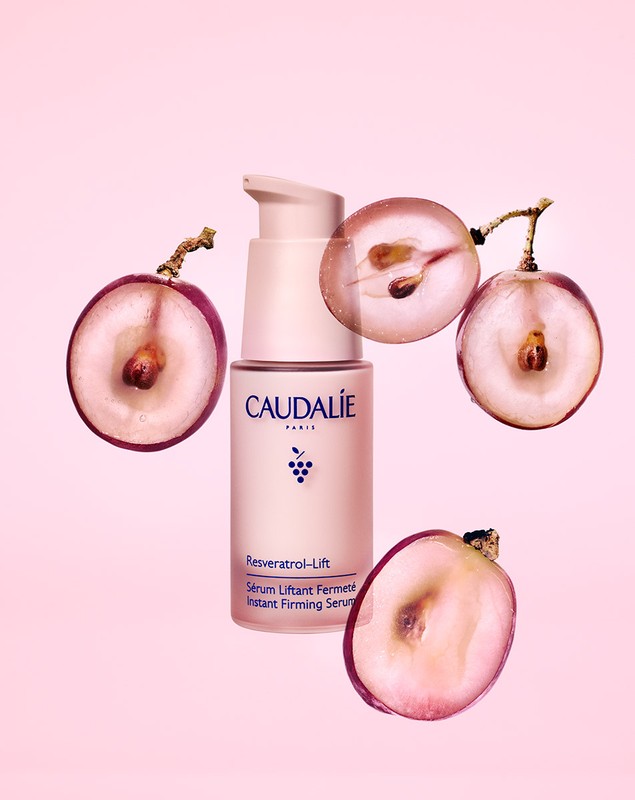 Caudalie Resveratrol Lift serum bottle with sliced grapes on a pink background, product shot for beauty or skincare advertising.