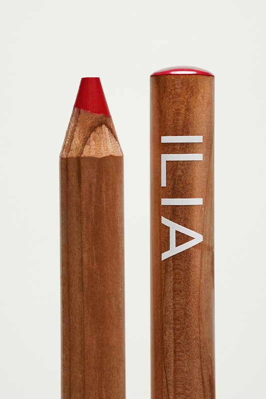 Close-up studio shot of a wooden lip pencil from Ilia Beauty with a red tip; product shot against a solid white background.