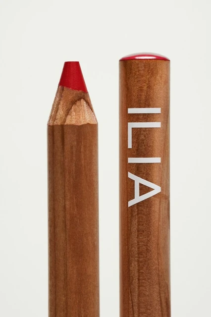 Close-up studio shot of a wooden lip pencil from Ilia Beauty with a red tip; product shot against a solid white background.
