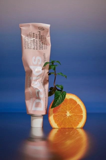Close-up shot of a tube of Dads toothpaste beside an orange half and a small green sprig against a gradient blue background.