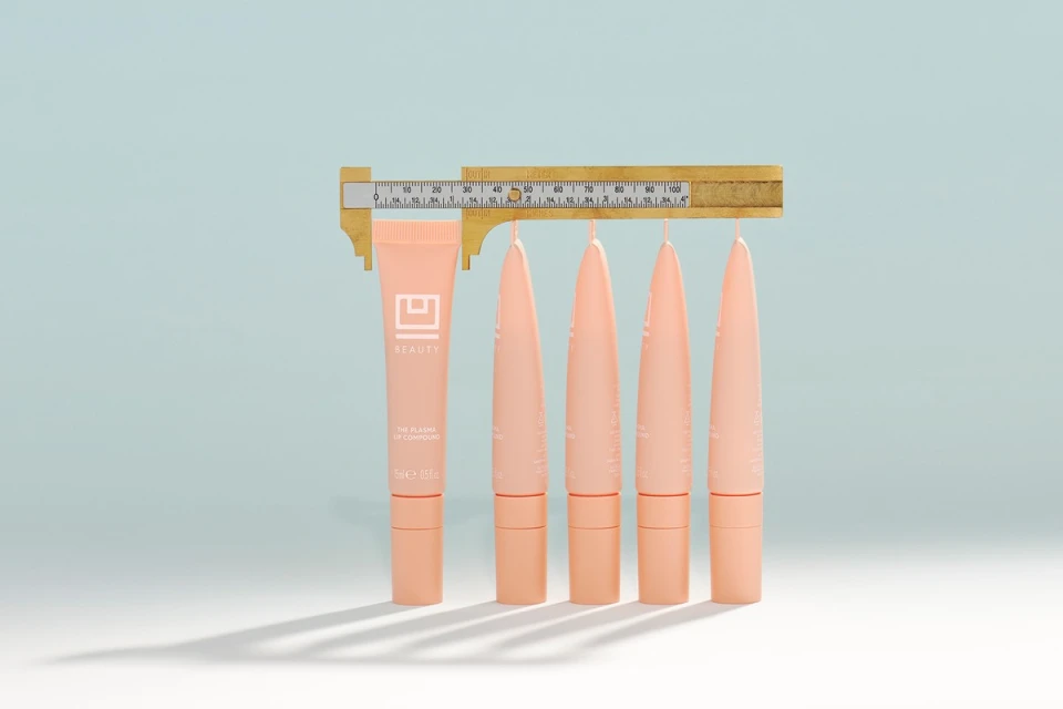 Peach lip compound tubes, measured by a gold-colored caliper on a gradient light blue background in a beauty product studio shot.