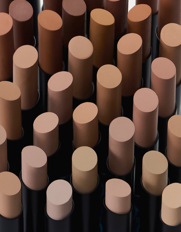 Close-up of multiple foundation makeup sticks in varying neutral skin tones, arranged in neat rows, shot in a studio setting.