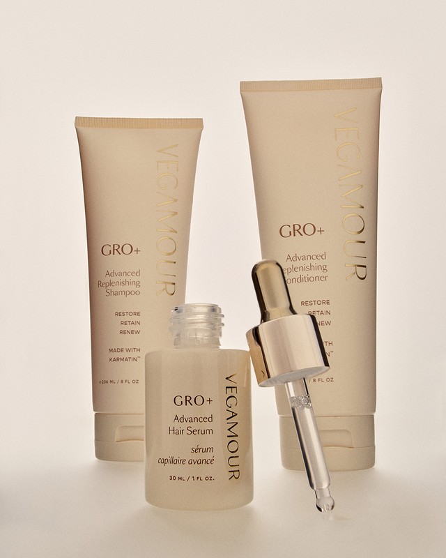 Three Vegamour brand hair products including shampoo, conditioner, and hair serum sit on a white surface in a studio setting.