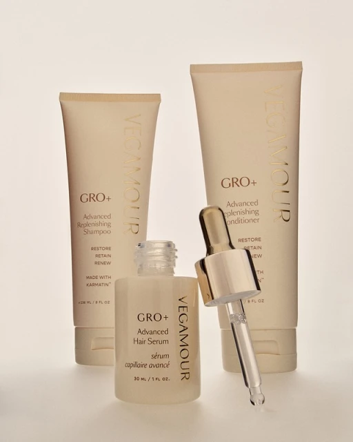 Three Vegamour brand hair products including shampoo, conditioner, and hair serum sit on a white surface in a studio setting.