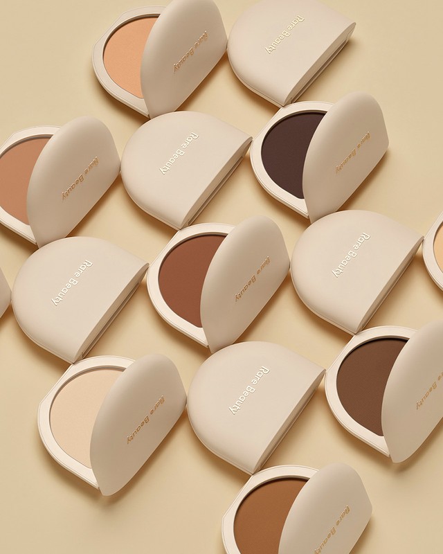 Close-up of multiple Rare Beauty compacts with open lids revealing different shades of makeup powder, arranged on a beige surface.