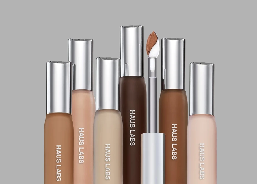 Several bottles of Haus Labs foundation in varying shades are arranged against a neutral gray backdrop. Beauty product flat lay.