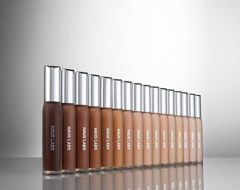Cosmetics display of Haus Labs foundation bottles in gradient shades, arranged in a row against a gradient gray studio backdrop.