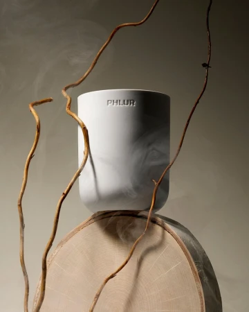Still life of a white cylindrical container with the word PHLUR on it, resting on a round slice of wood, accented by winding branches.