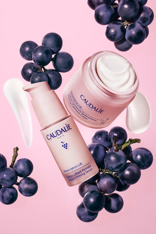 Caudalie skincare products featuring a cream jar, serum bottle, and fresh grapes arranged against a soft pink background in a studio setting.