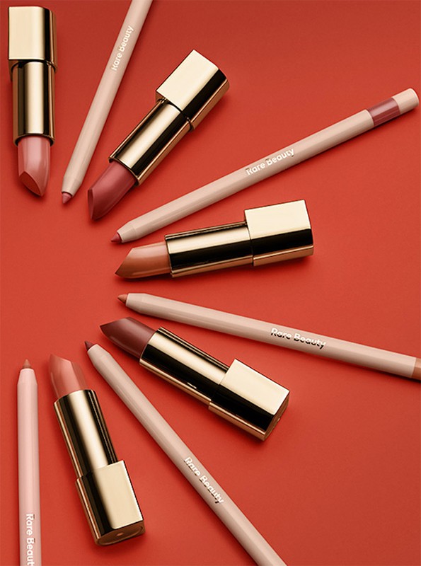 Still-life shot of several Rare Beauty lipsticks and lip pencils arranged diagonally on a red background, capturing makeup essentials.
