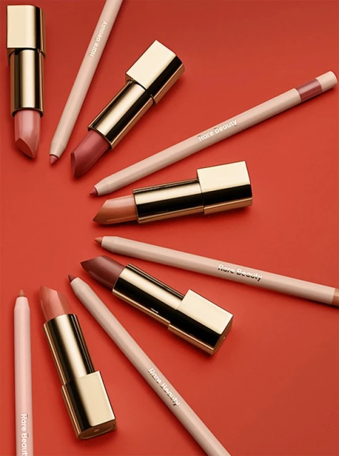 Still-life shot of several Rare Beauty lipsticks and lip pencils arranged diagonally on a red background, capturing makeup essentials.