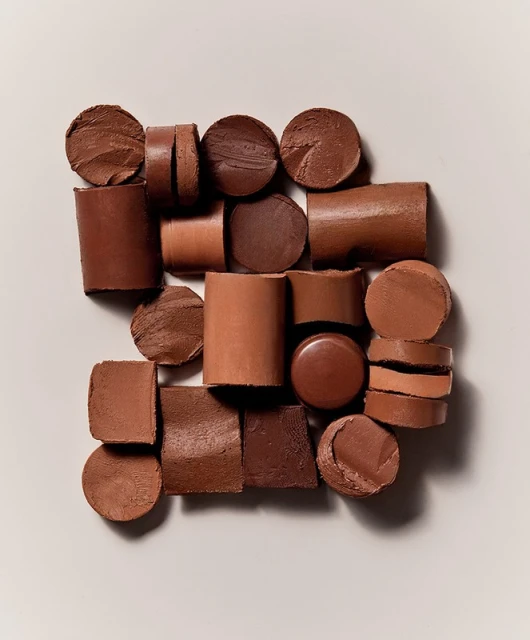 Geometric arrangement of cylindrical and circular chocolate cosmetic products of varying brown hues, set against a soft, neutral background.