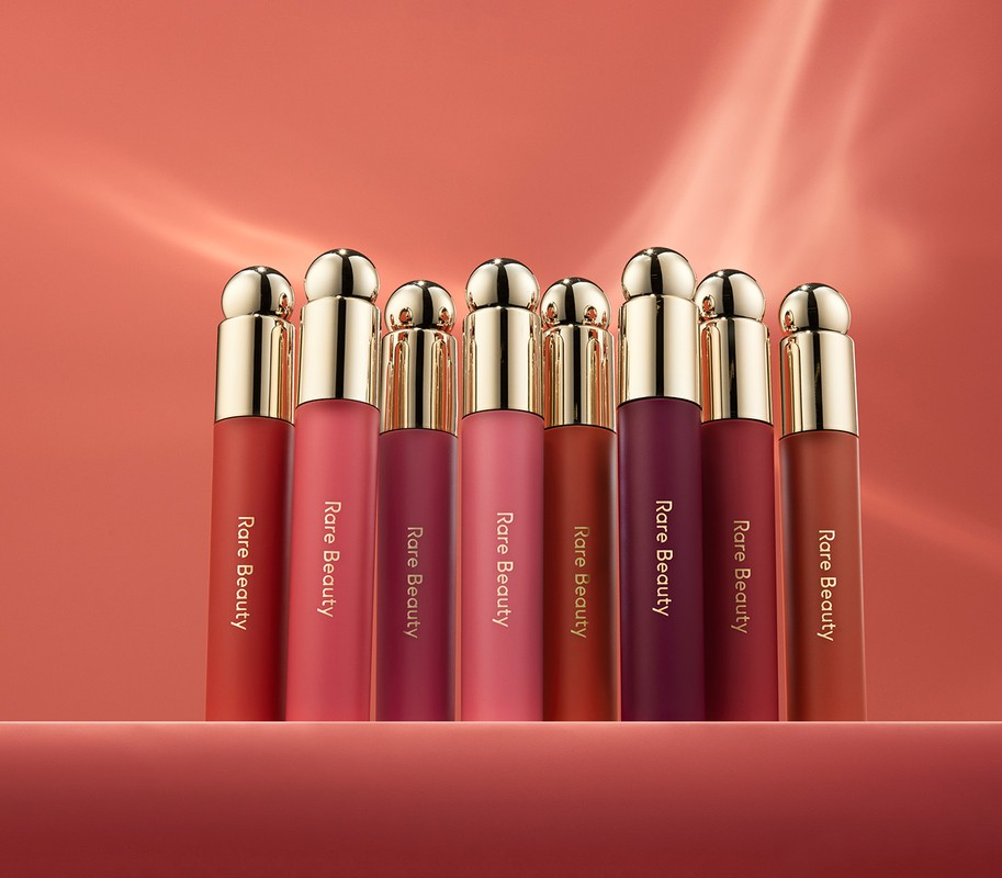 Eight Rare Beauty lipsticks of varying colors stand in a row on a surface against a peach-colored background with light reflections.