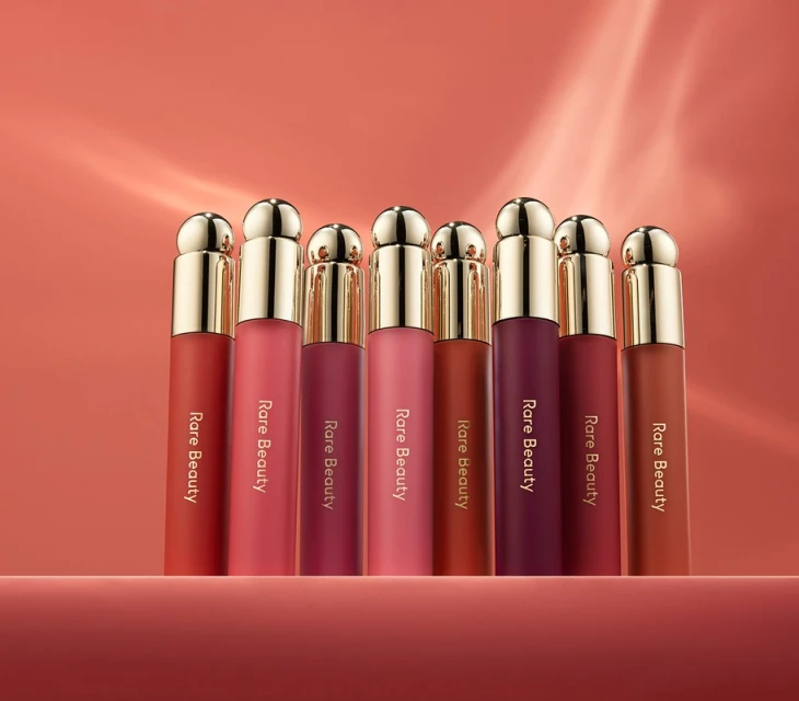 Eight Rare Beauty lipsticks of varying colors stand in a row on a surface against a peach-colored background with light reflections.