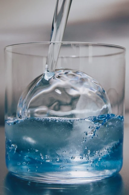 Water being poured into a clear glass filled with ice, creating bubbles and refraction, on a neutral-colored surface indoors.