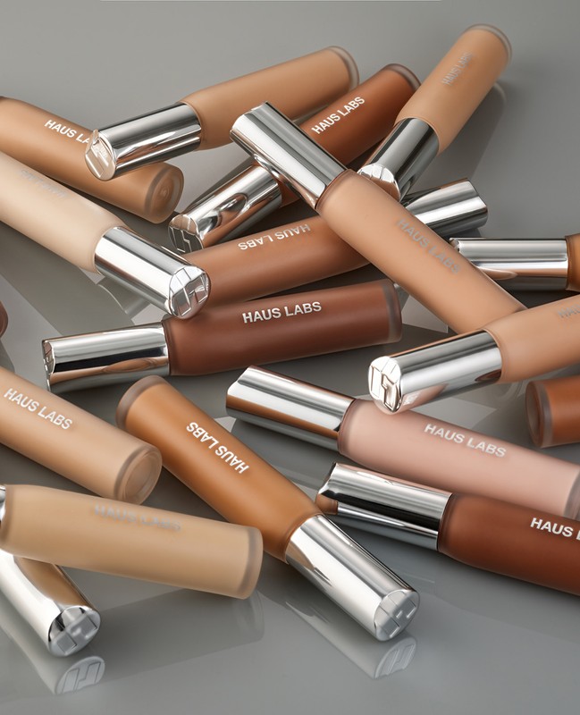 Cosmetic product tubes of Haus Labs foundation in varying skin tones, arranged on a gray surface, showcasing a range of shades.